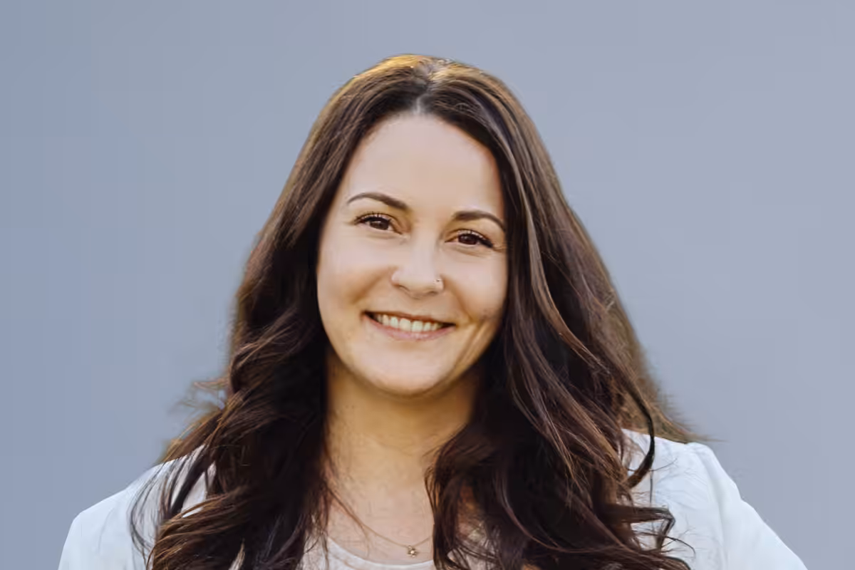 Smiling woman with long brown hair wearing a light jacket against a gray background.